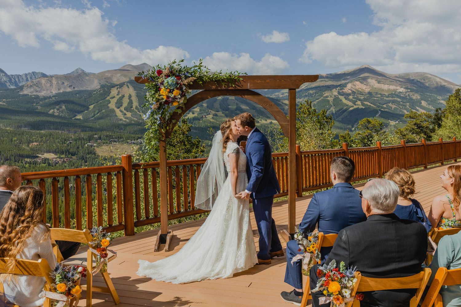 A mountain wedding ceremony takes place on a wooden deck with a floral arch and stunning Colorado peaks in the background.