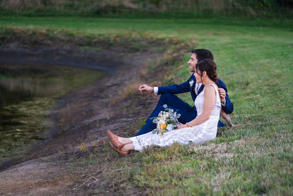A bride and groom sit casually on the banks of a small pond at a backyard wedding event in South Haven, Michigan.
