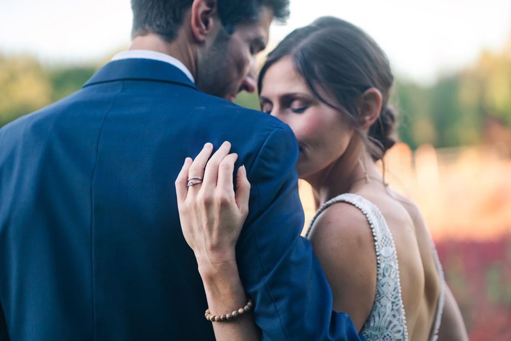 The bride's new wedding ring is displayed on her hand on his shoulder as they snuggle during golden hour.