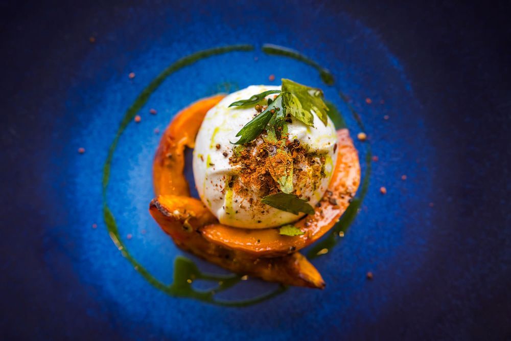 Artistic plating of burrata topped with herbs and spices, paired with roasted squash, drizzled with green oil on a vibrant blue plate.