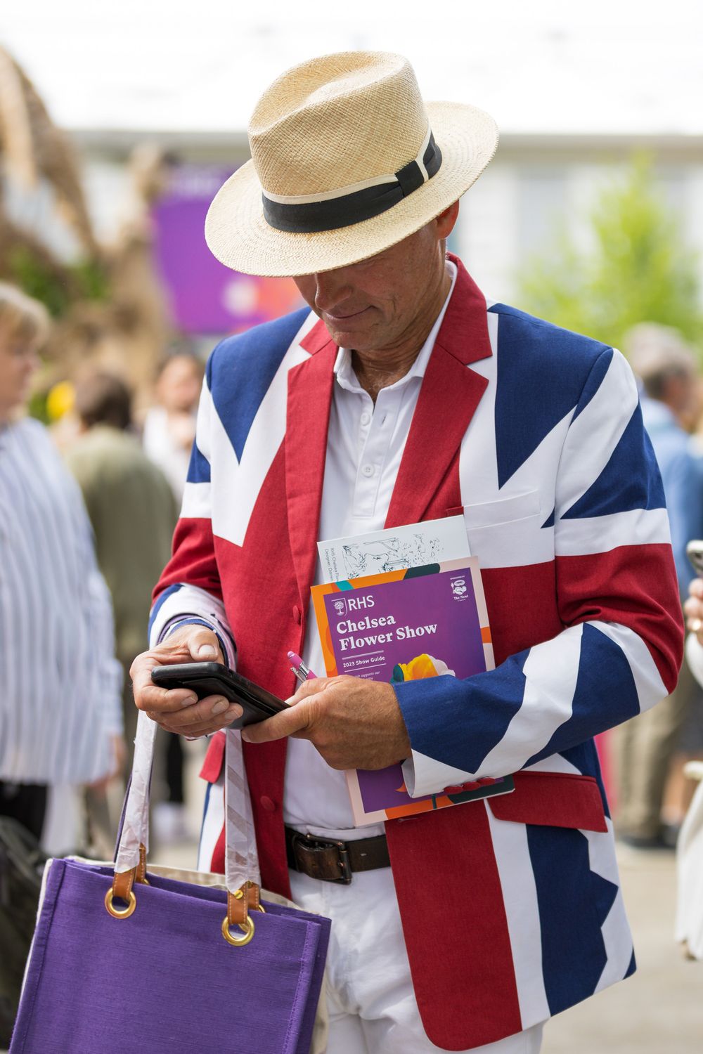 A spectator in a Union Jack blazer and straw hat checks their phone while holding a purple bag at a sporting event.