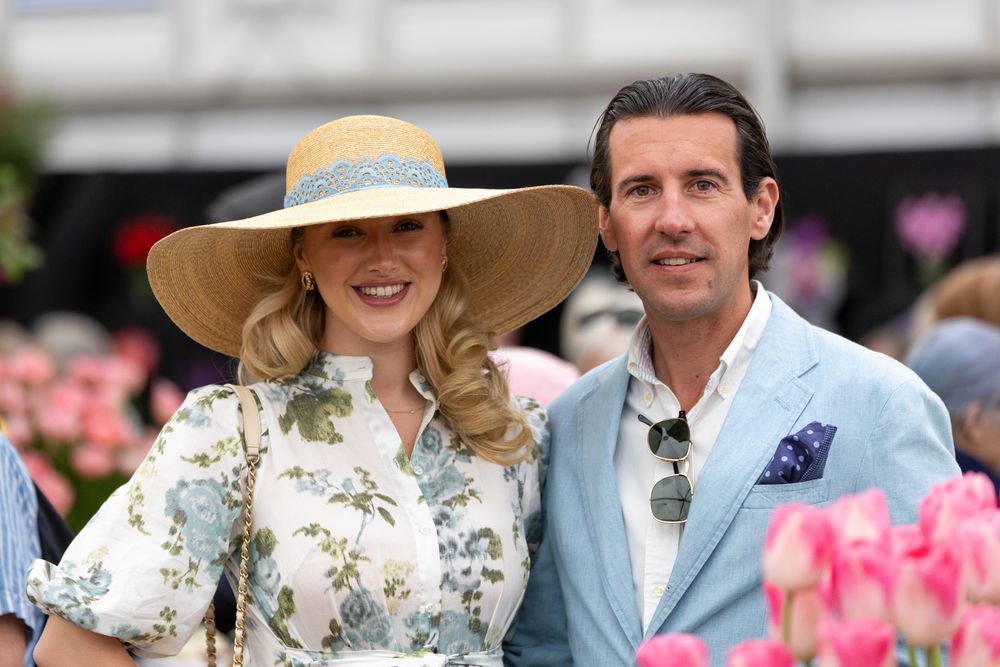 Couple at spring event with woman in floral dress and wide-brimmed hat and man wearing light blue blazer near pink flowers.
