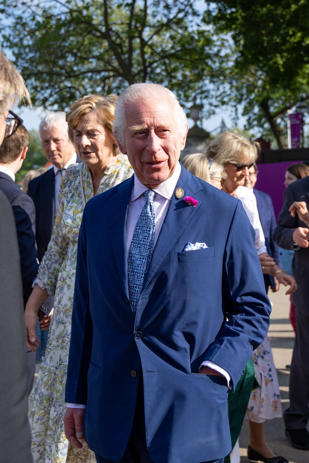 A person in a navy blue suit and tie with a red poppy pin stands outdoors among a group of people on a sunny day.