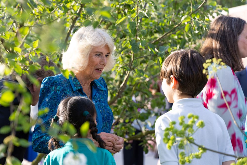 A person wearing a blue dress interacts with children in a lush garden setting surrounded by green foliage.
