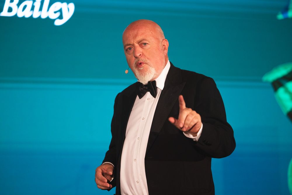 Comedian Bill Bailey in a black tuxedo and bow tie gestures while speaking on stage with teal lighting in the background.