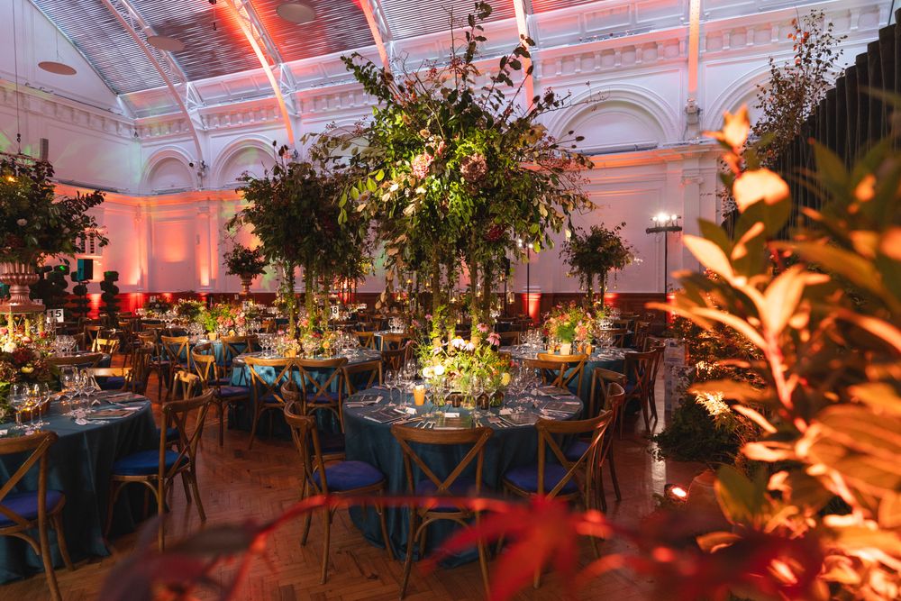Elegant wedding reception venue with dramatic floral centerpieces, blue tablecloths, and pink ambient lighting in ornate ballroom.