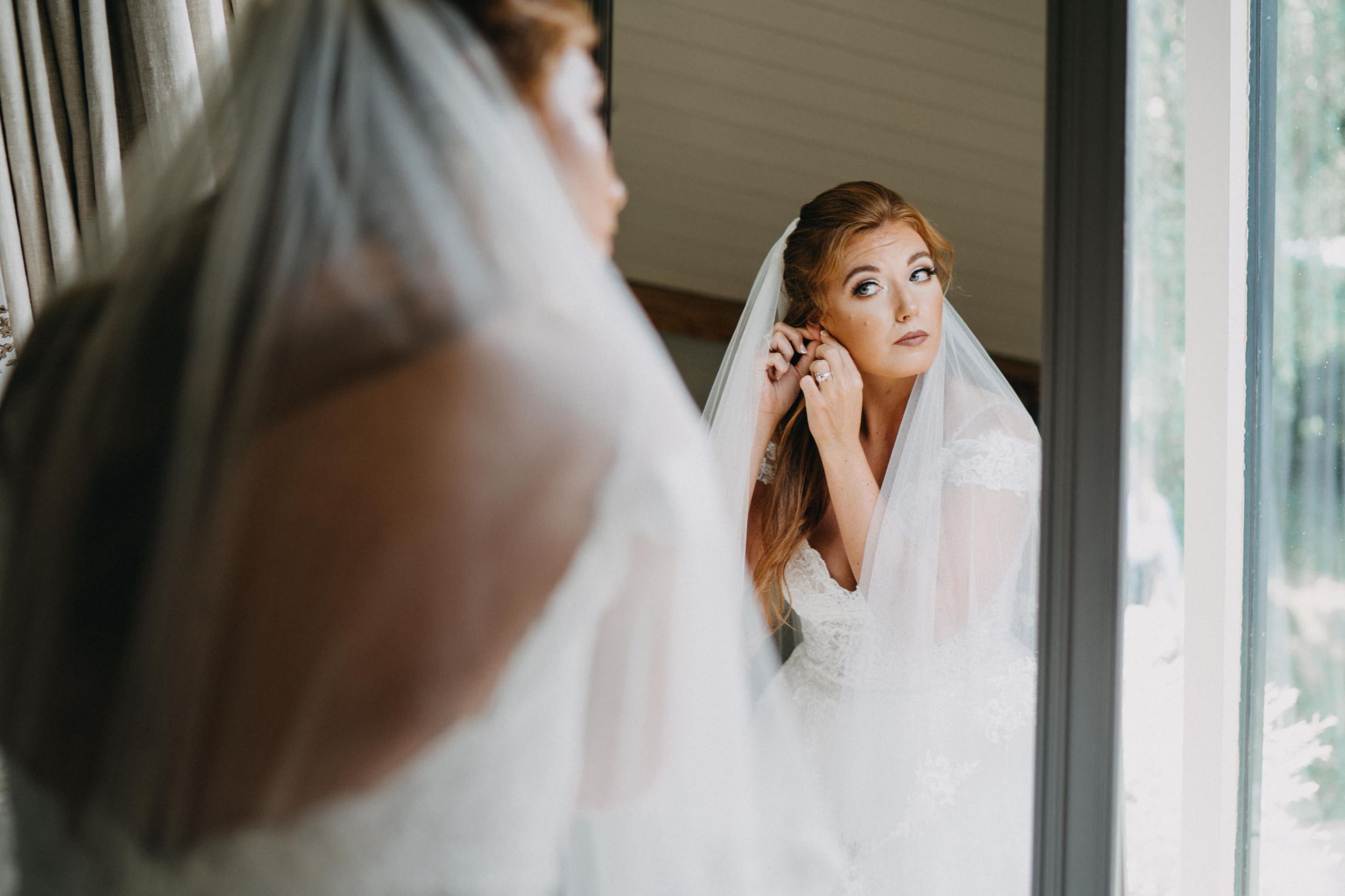 Bride putting on earrings while looking in mirror during wedding preparations.