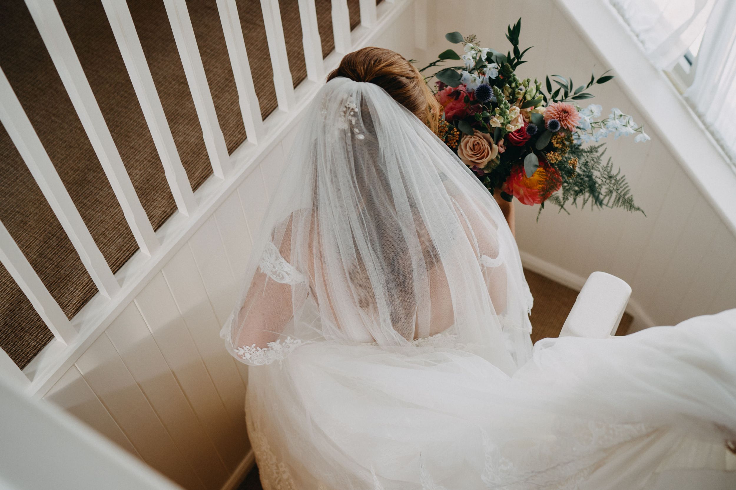 A bride in a white wedding dress and veil sits on stairs next to a colorful floral arrangement.