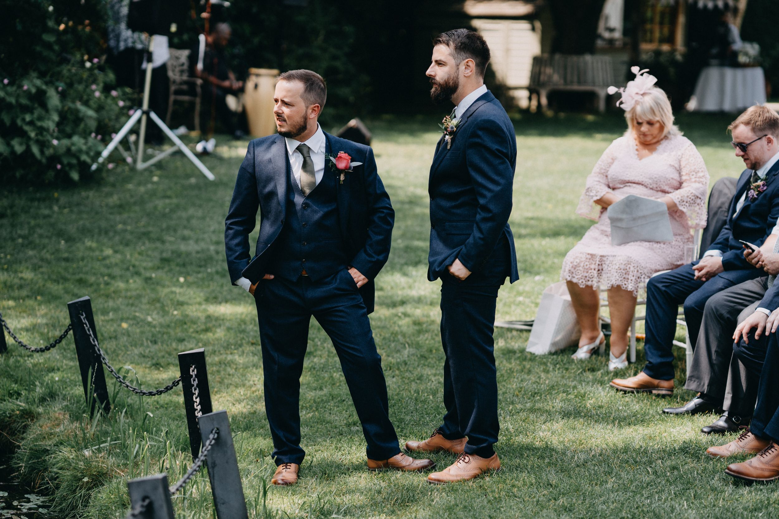 Wedding guests in formal attire stand on grass during an outdoor ceremony.