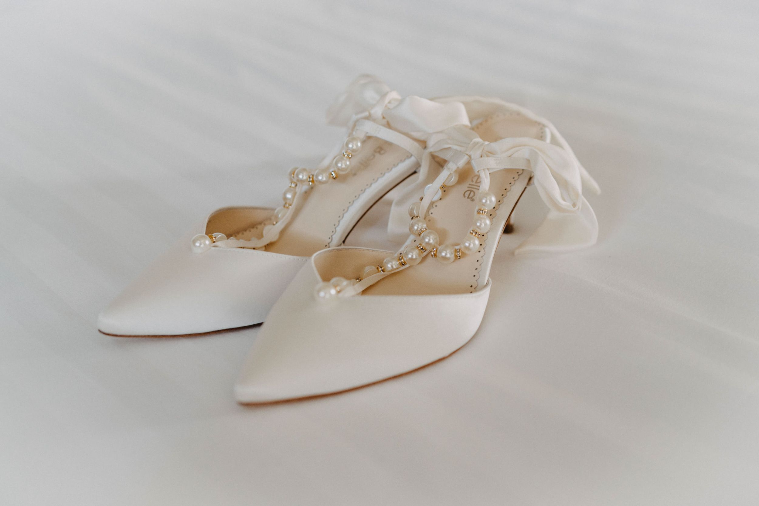 Elegant white bridal shoes with pearl straps on light fabric background.