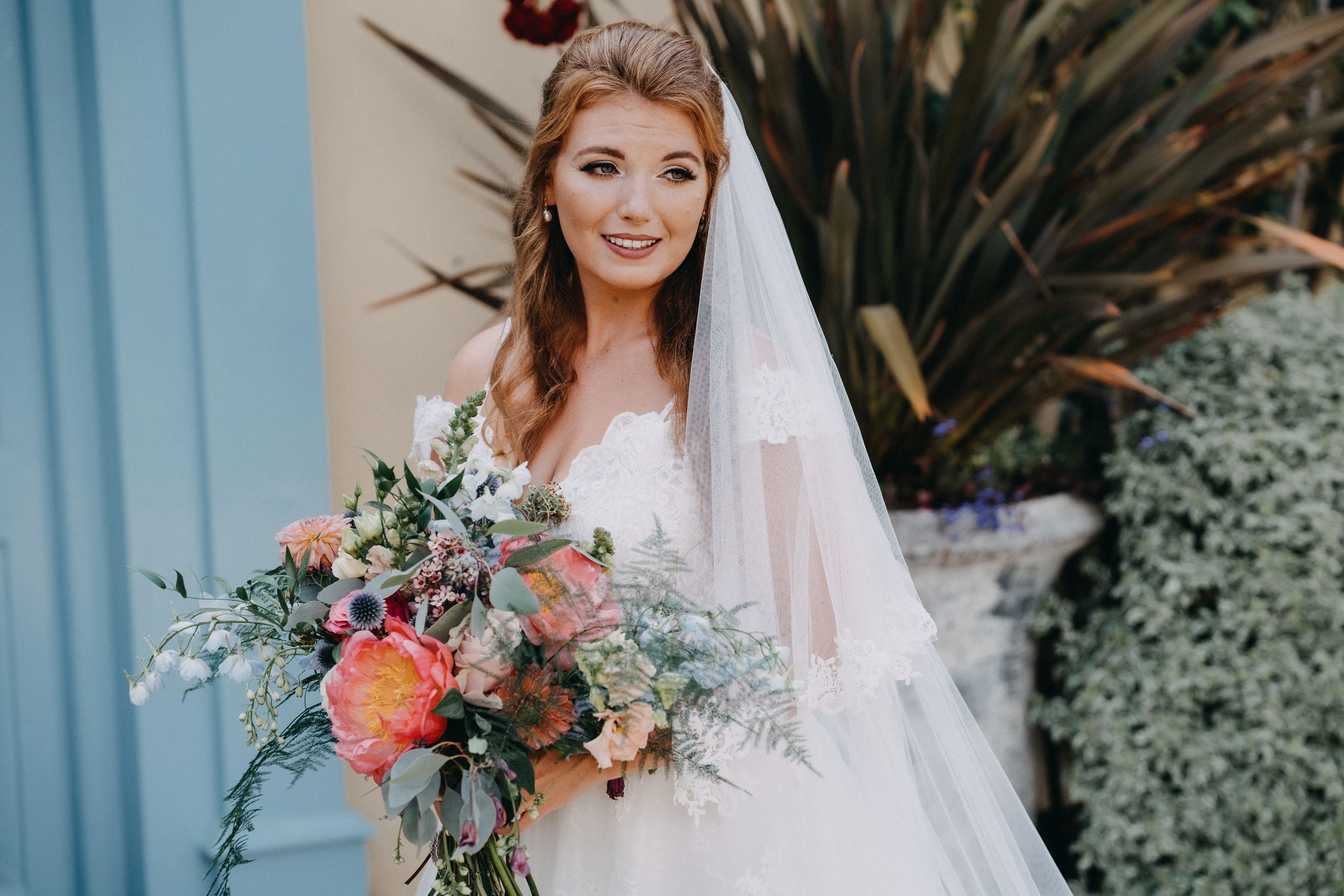 Bridal portrait with coral and peach bouquet against blue wall and tropical plants.