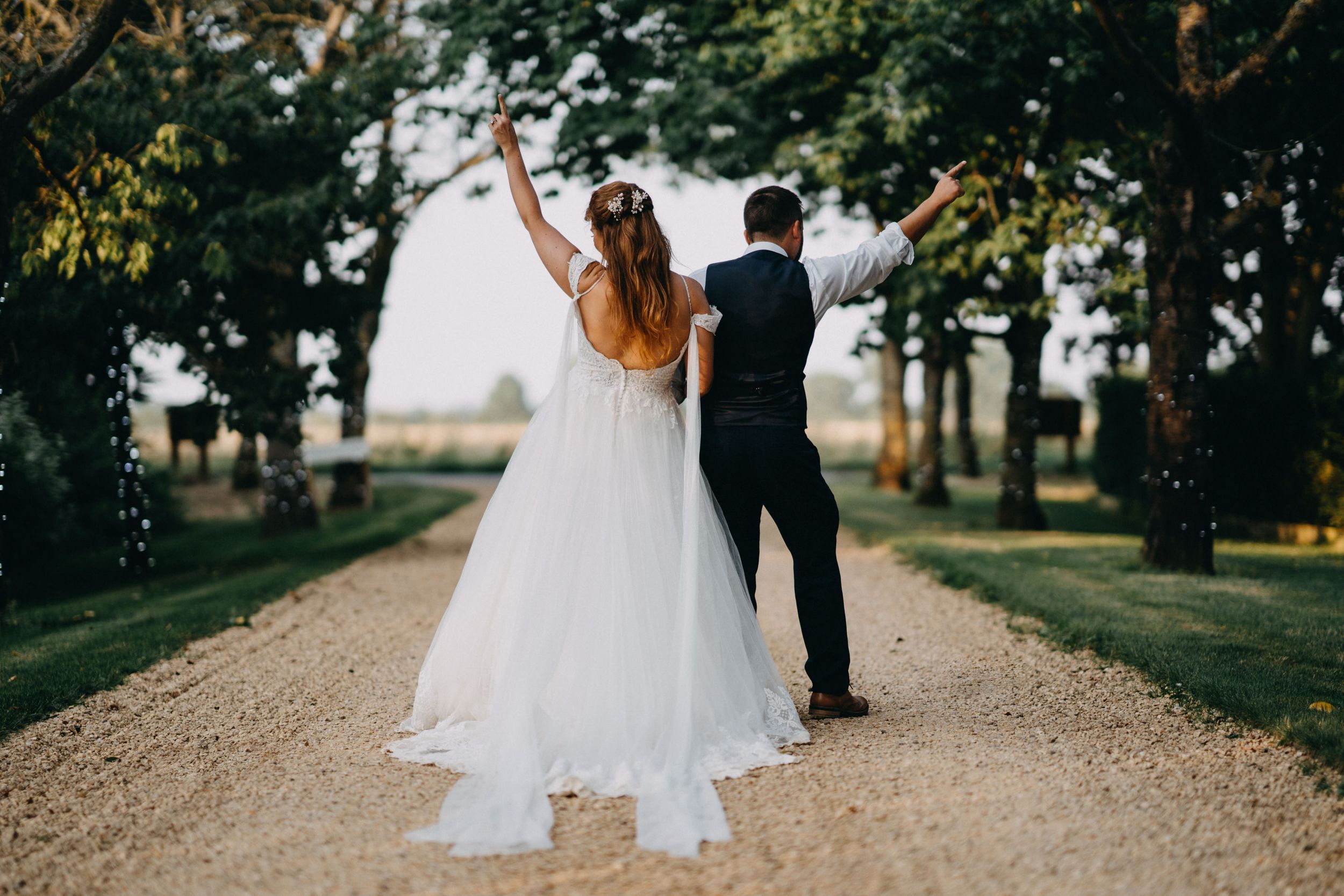 Newlyweds celebrate with raised arms on a tree-lined gravel path at sunset.