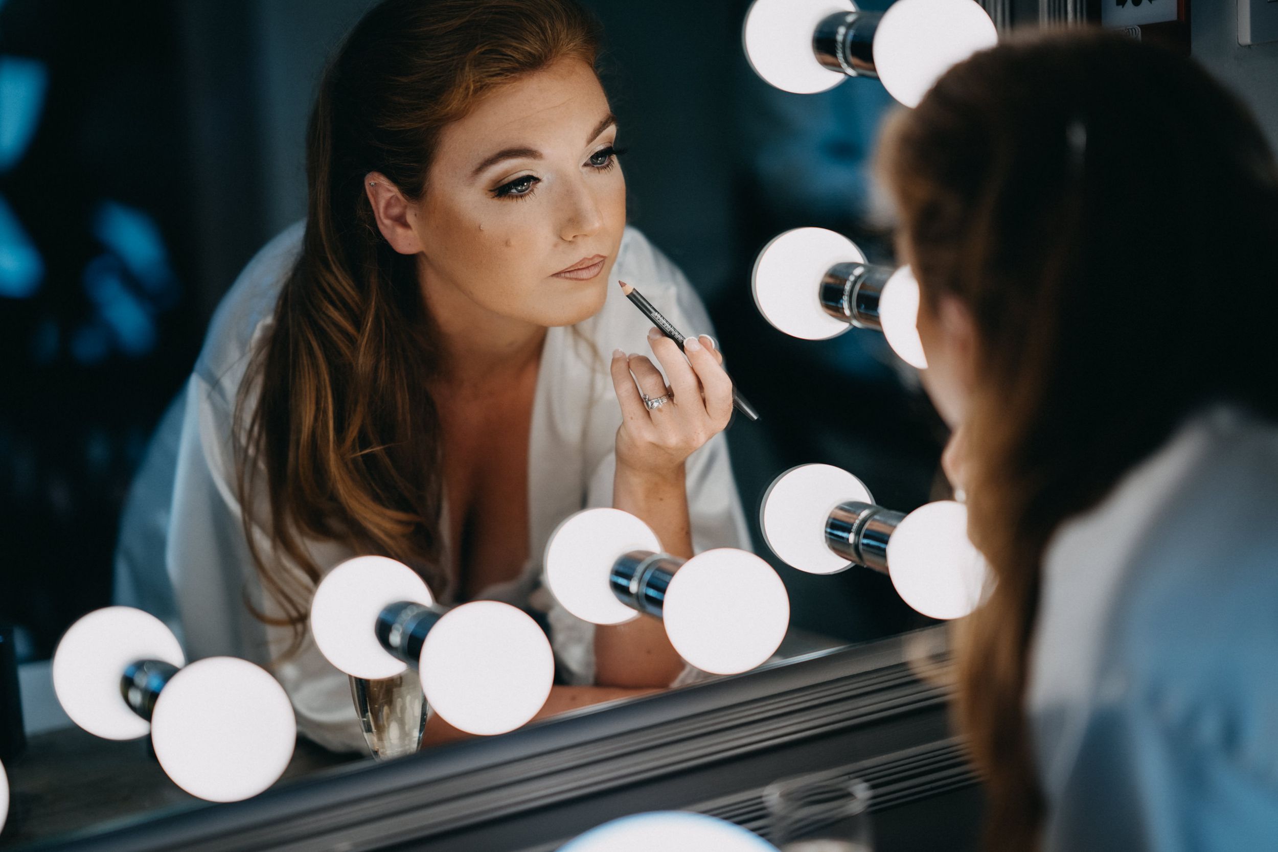 Woman applies makeup in front of a vanity mirror with round bulbs creating a glamorous atmosphere.