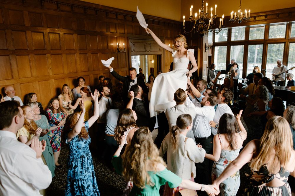 Wedding celebration with guests cheering as a person in a white dress is lifted high into the air during a reception.