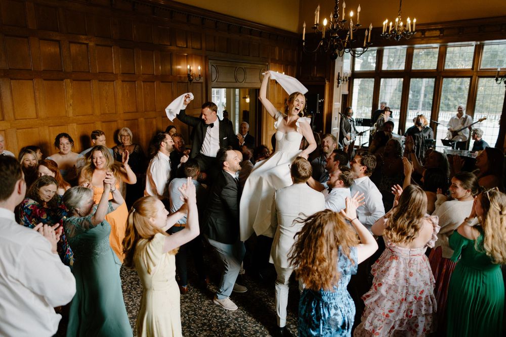 Wedding guests dancing and celebrating at an elegant reception inside a wood-paneled ballroom with chandelier lighting.