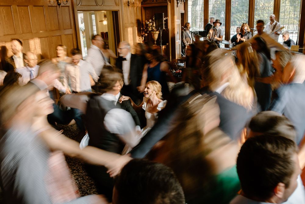 Motion blurred crowd dancing together at a celebration in an elegant wood-paneled hall with large windows.