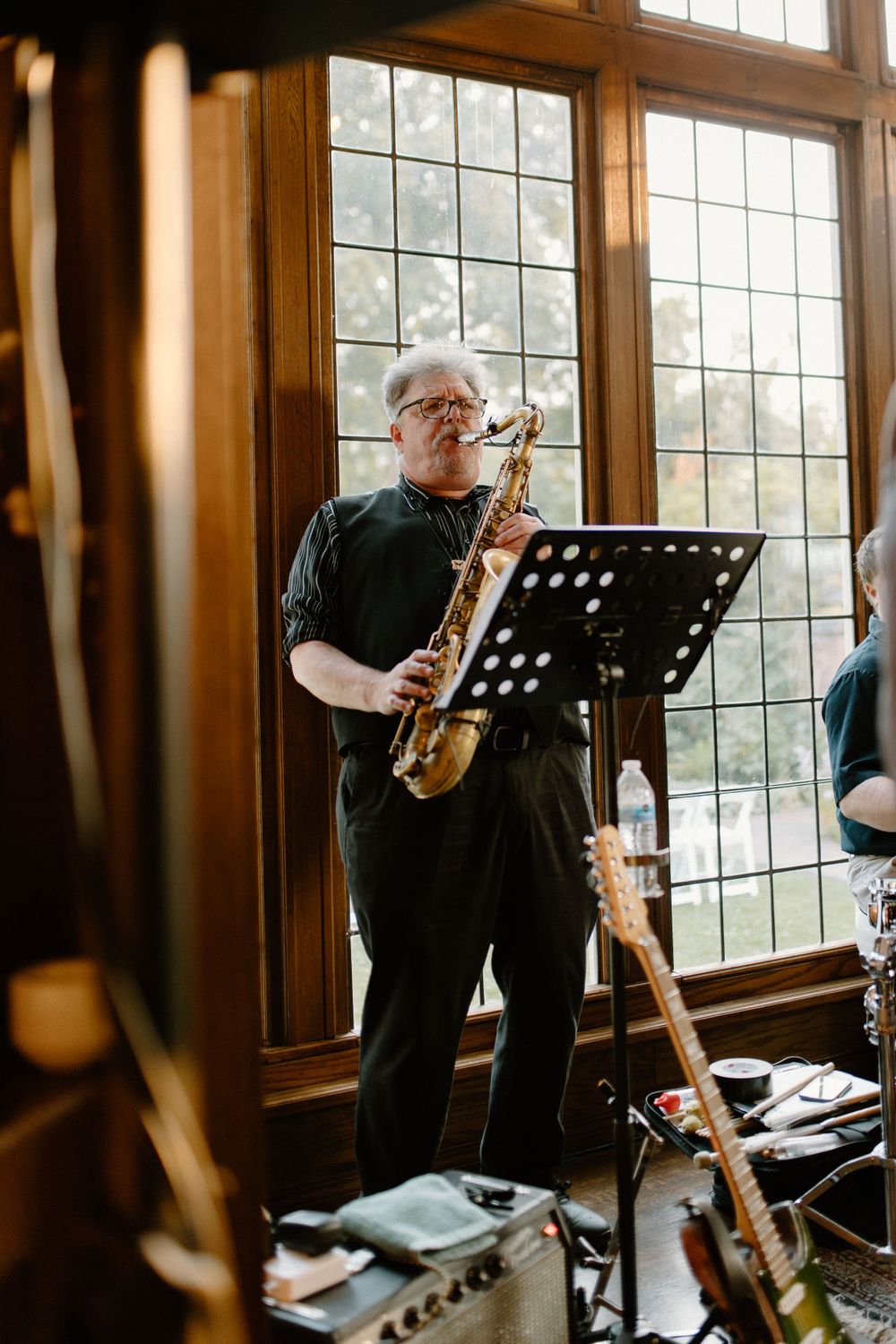 Musician plays saxophone near large leaded glass windows in warm wood-paneled room with music stand.