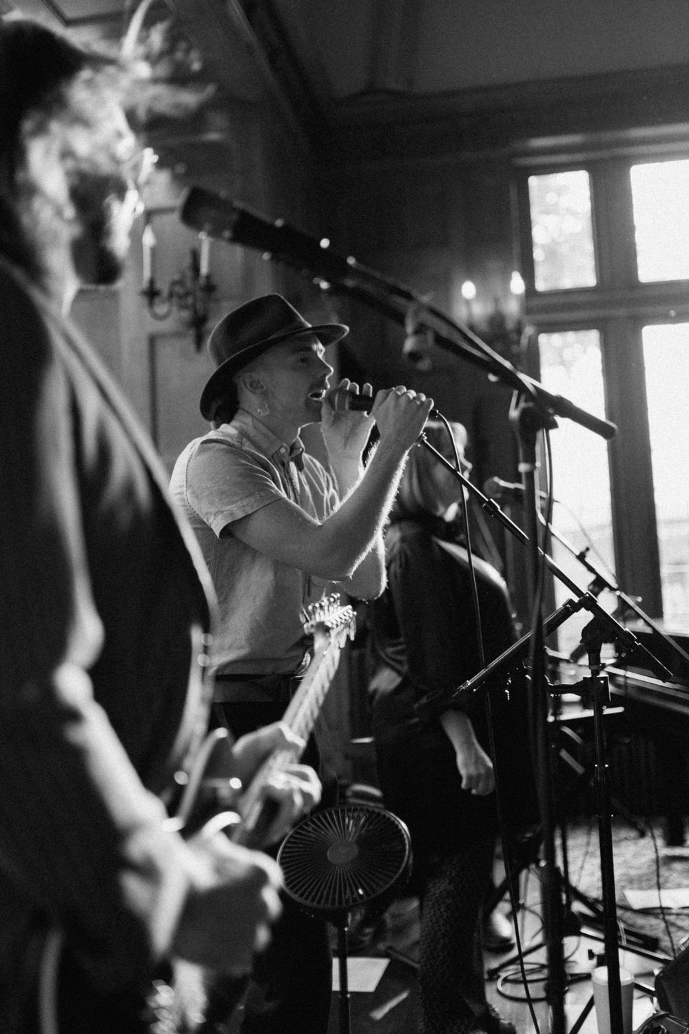 Black and white concert photo of a musician performing with a microphone on stage with band members visible in shadows.