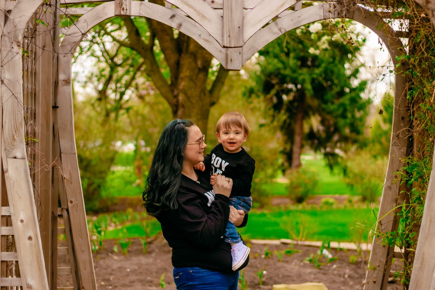 Two people share a sweet moment together under a rustic wooden archway in a lush garden setting.