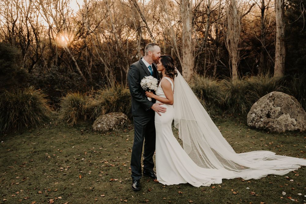Romantic sunset wedding portrait with a flowing white veil against a golden woodland backdrop.