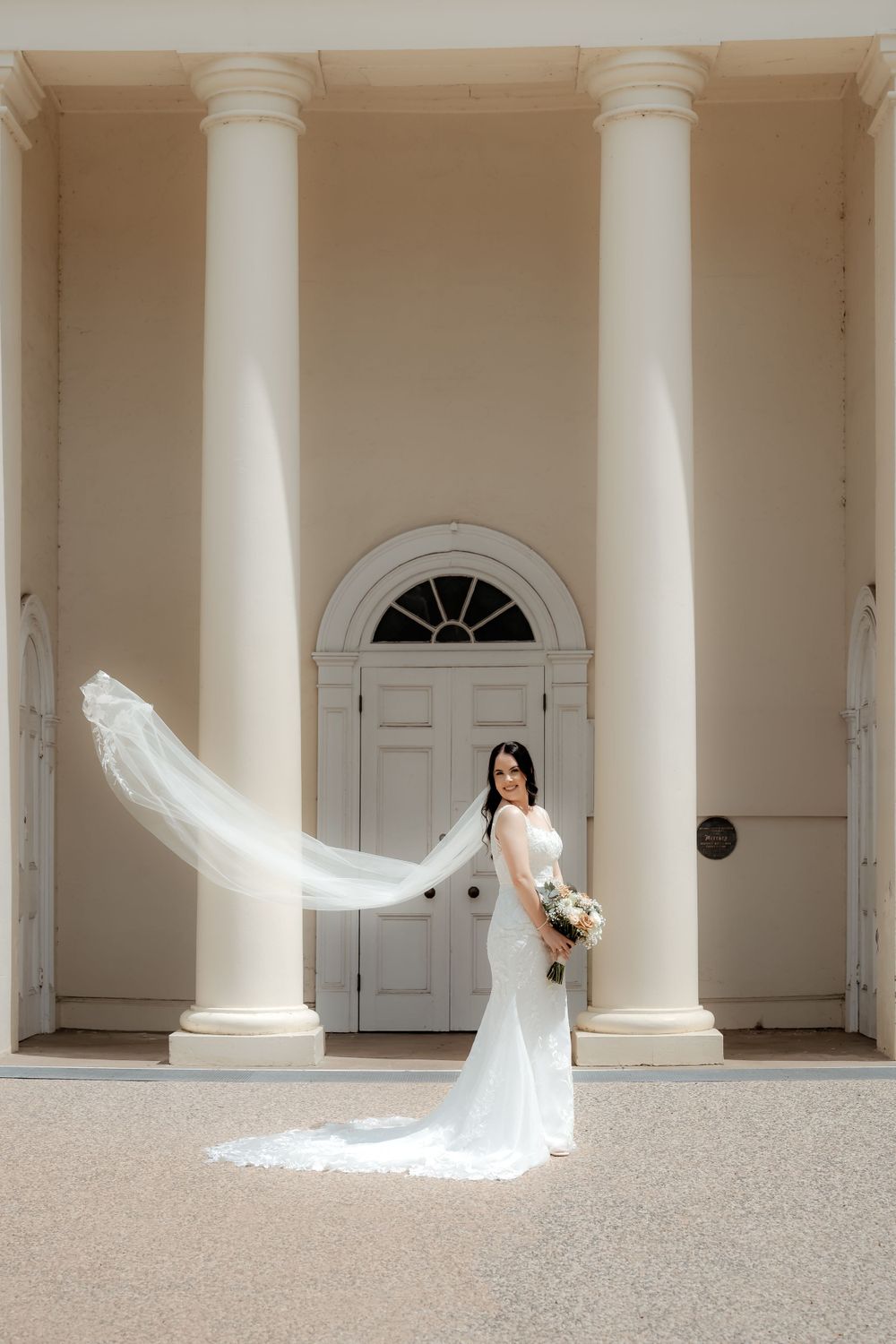 Elegant bridal portrait with flowing veil against white columns and door.