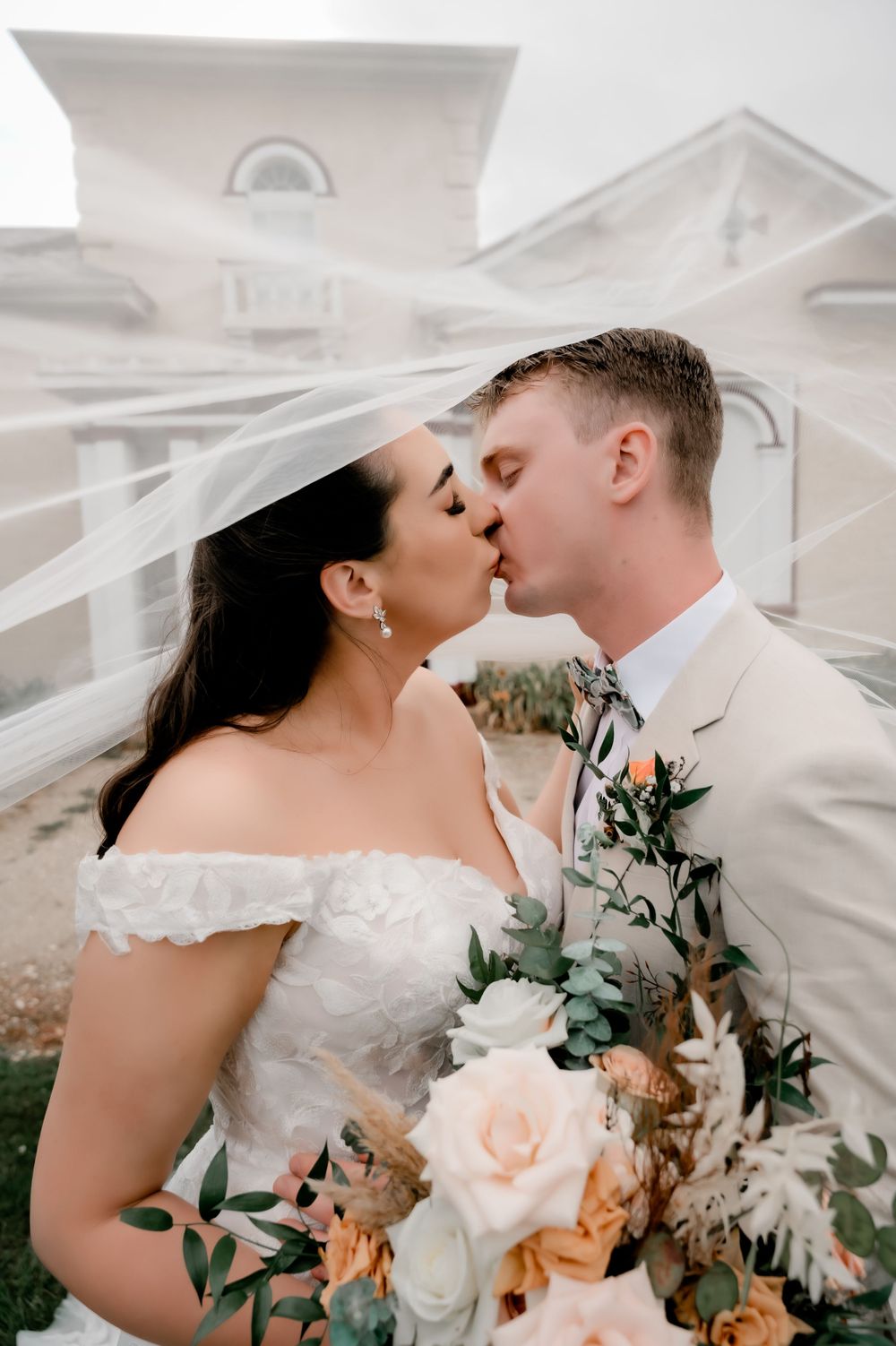 Romantic wedding kiss with flowing veil and peach floral bouquet.