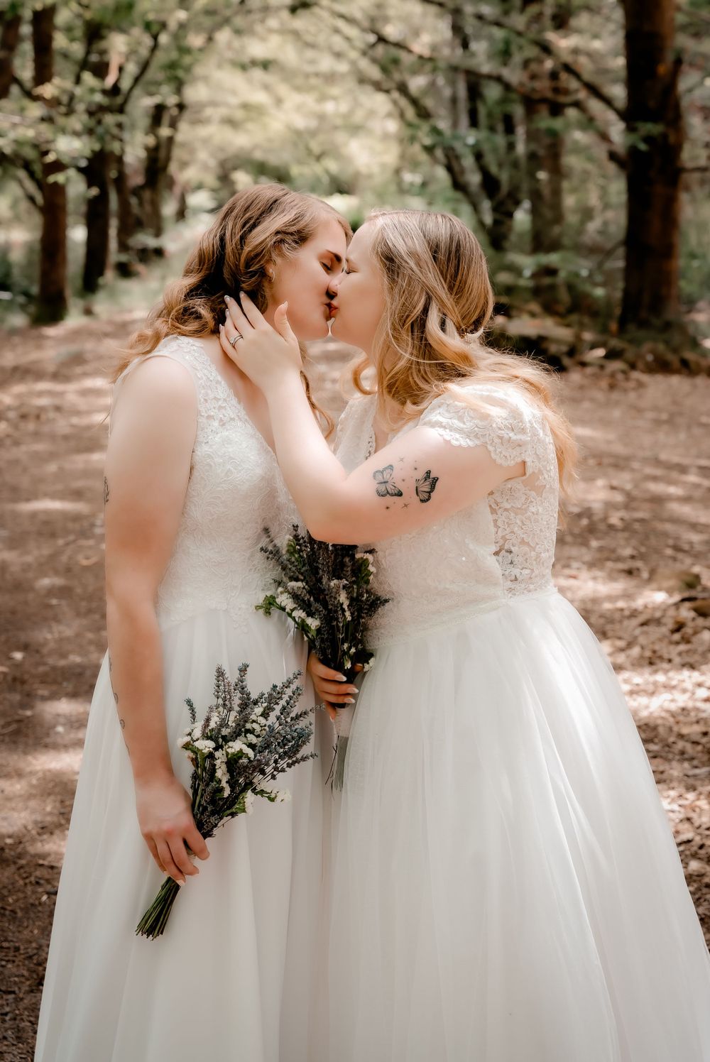 Two brides in white dresses share a kiss while holding bouquets in a forest setting.