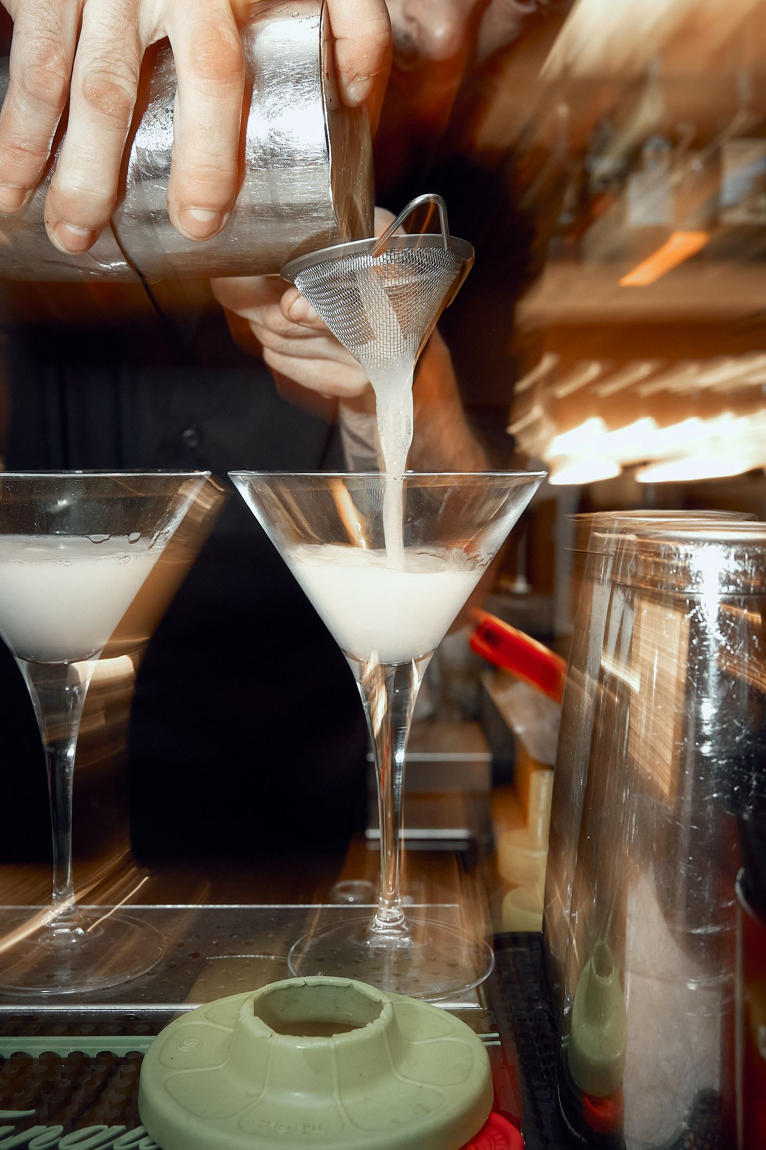 Bartender pouring a white cocktail through a strainer into martini glasses.