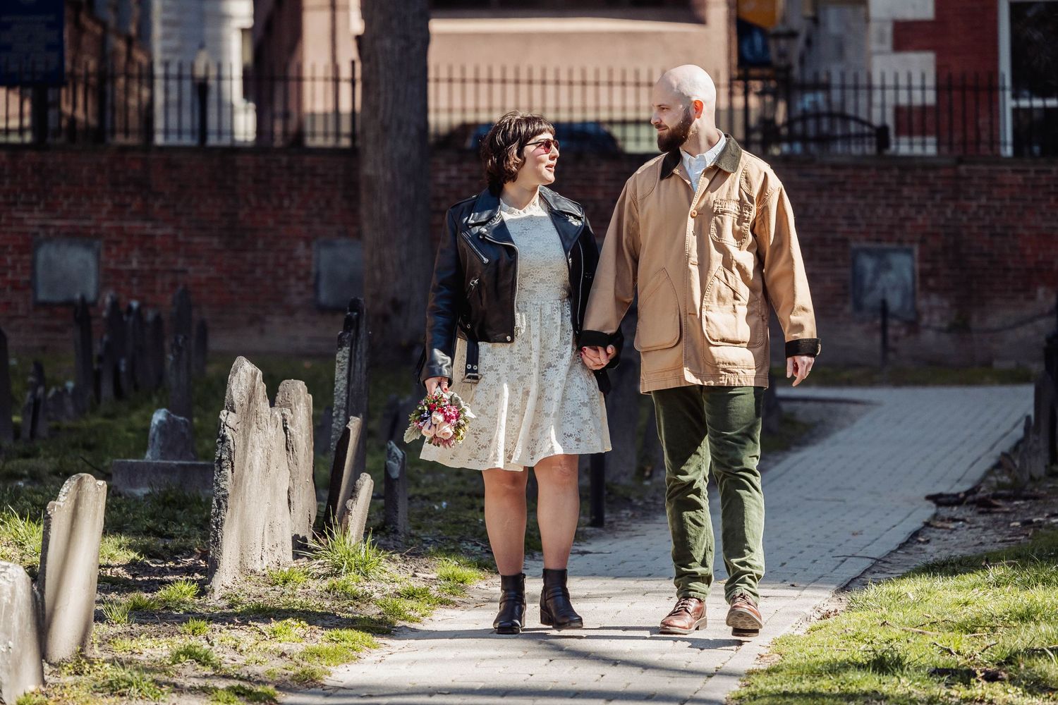 A couple strolls through a historic cemetery with brick buildings in the background.