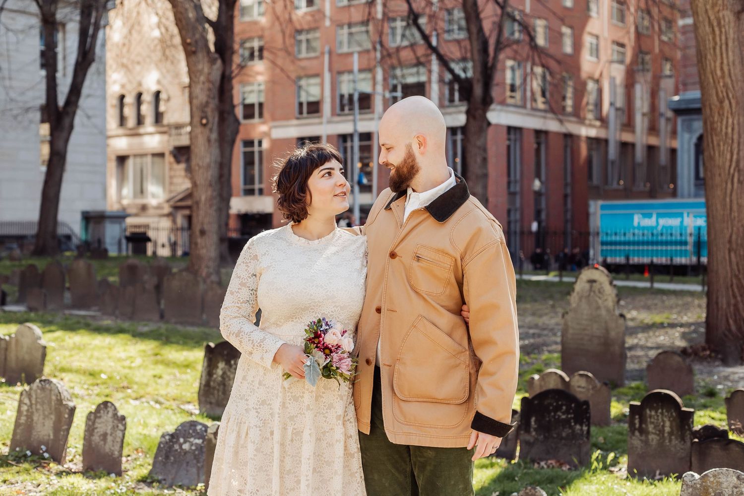A couple poses in a cemetery garden with historic brick buildings behind them.