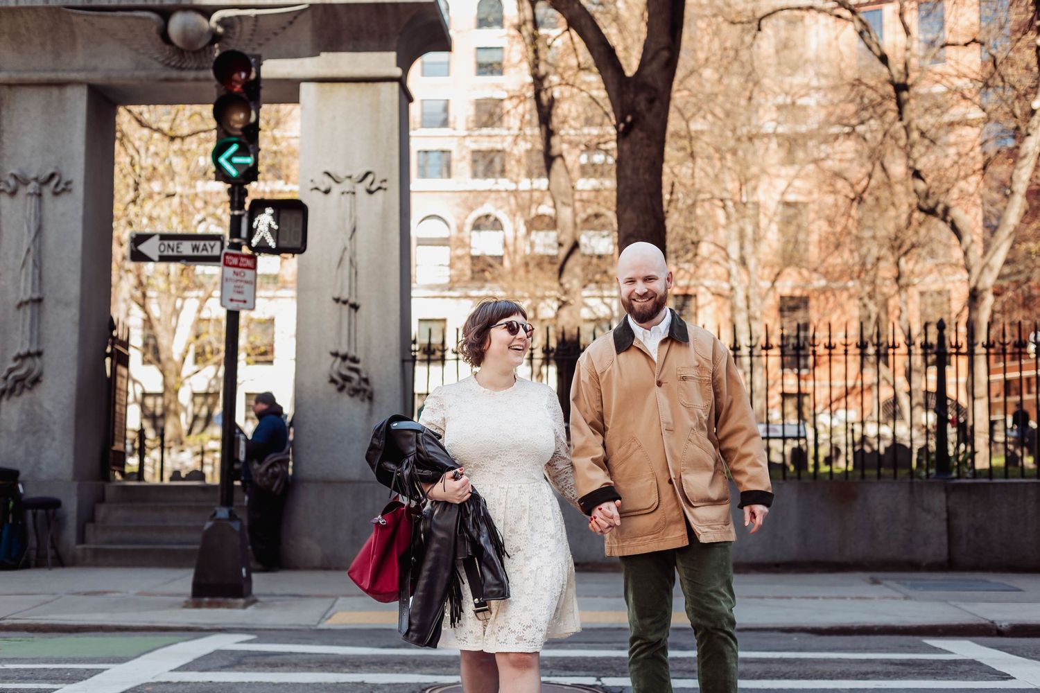 A couple stands at a city crosswalk wearing wedding attire.