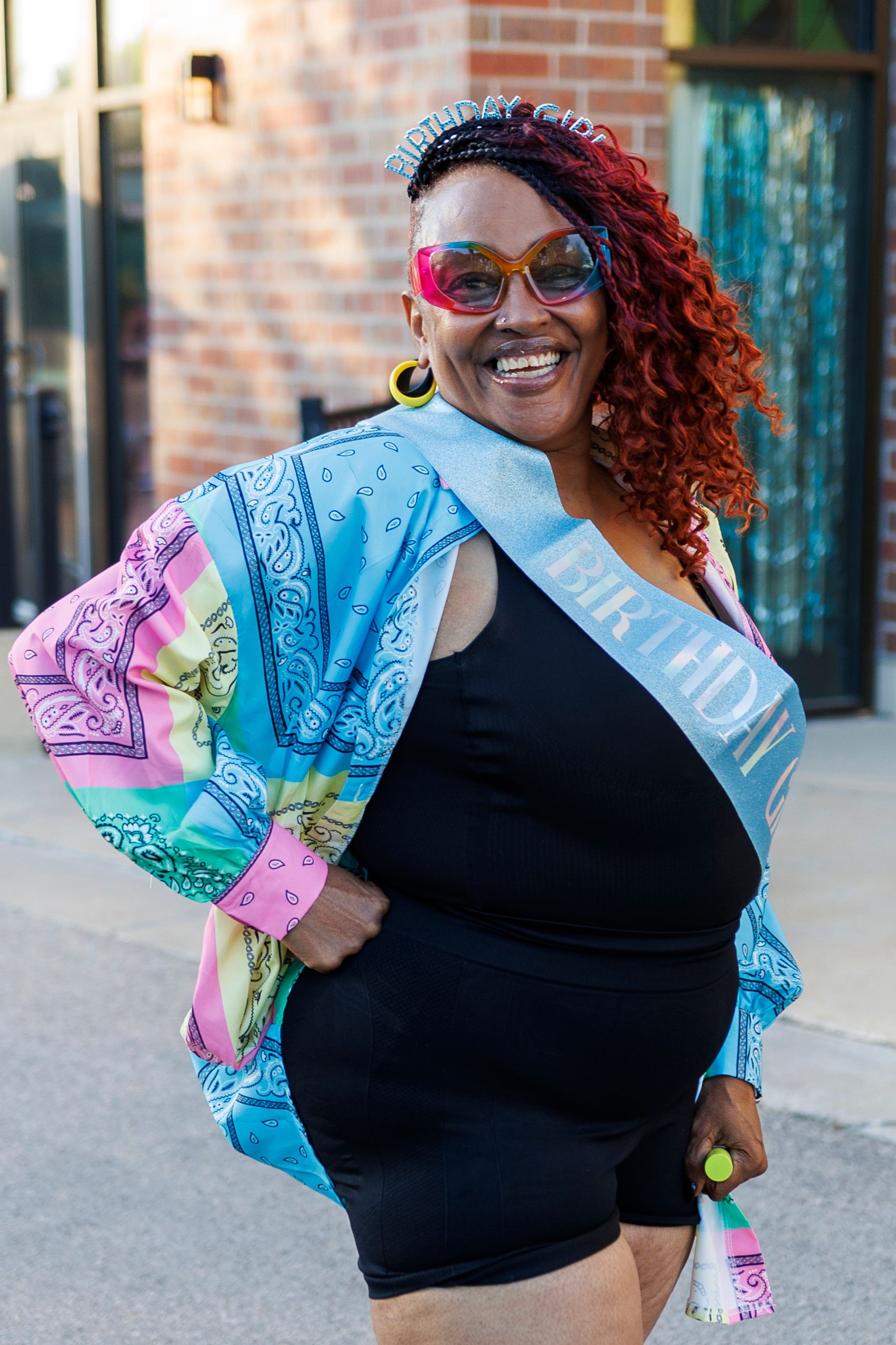 Person wearing colorful patterned jacket and black dress posing with bright sunglasses.