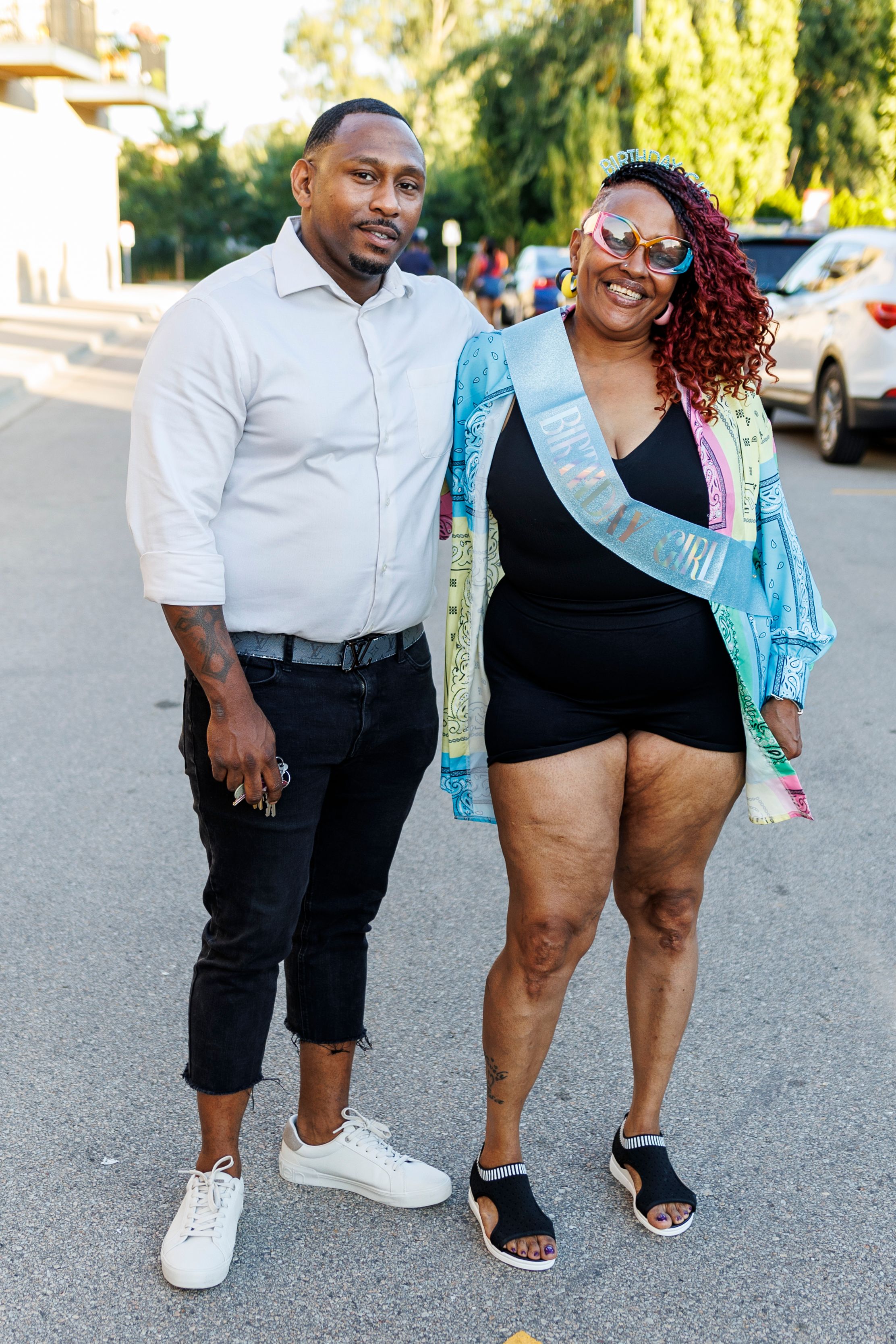 Couple posing on street, one wearing white shirt and jeans, other in black dress with blue sash.