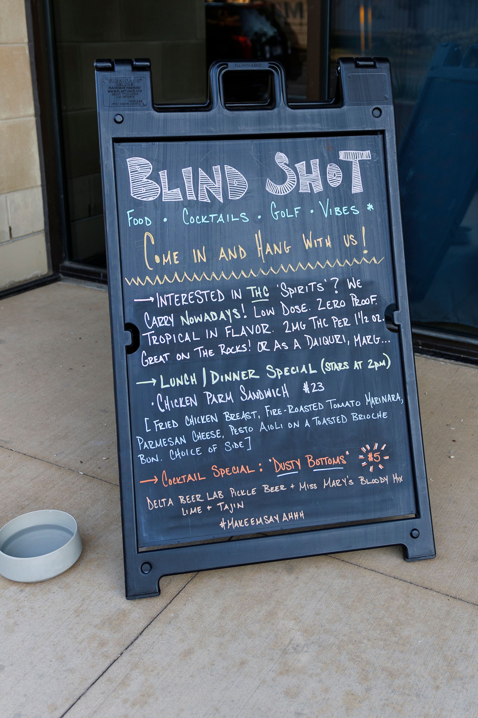 A chalkboard sign outside a business displays the menu and specials for 'Blind Shot' establishment.