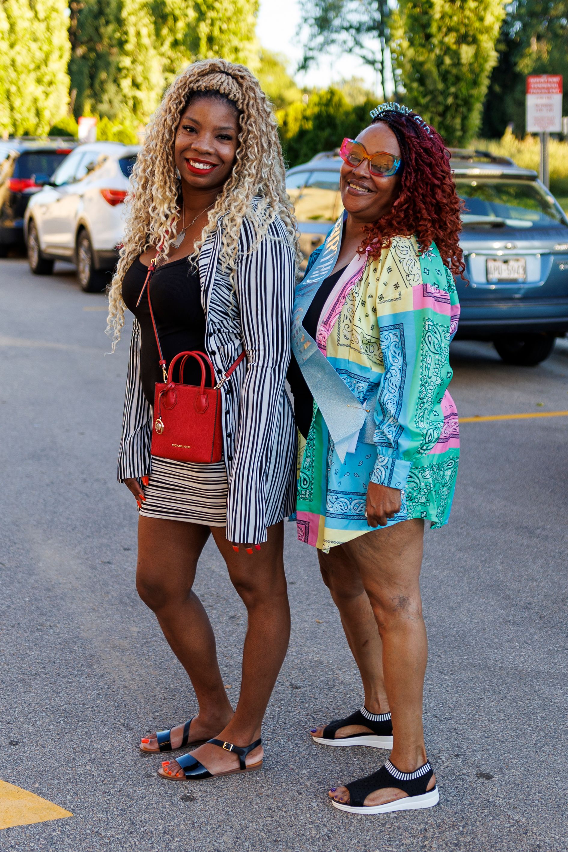Two fashionable friends pose together in a parking lot wearing colorful summer outfits and stylish accessories.