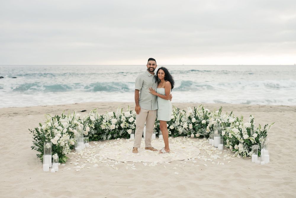 A romantic beach scene decorated with white floral arrangements in the sand as waves crash behind against an overcast sky.