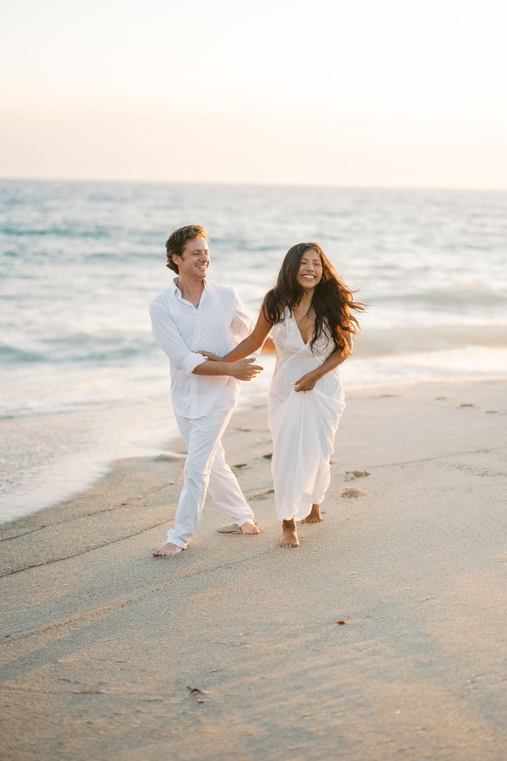A couple in white outfits playfully runs along a sandy beach at sunset.