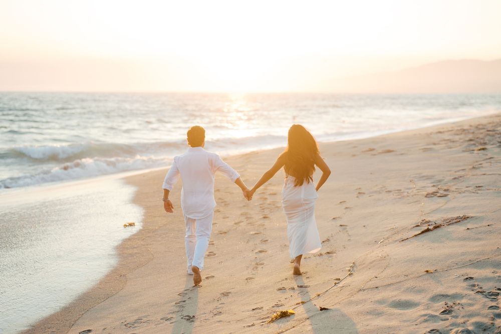 Two people in white clothing hold hands while walking along the beach during golden hour.