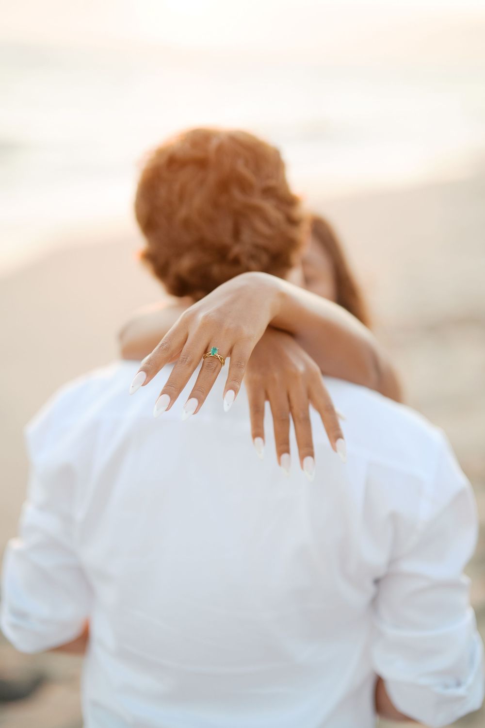 A close-up shot shows hands with an engagement ring resting on a white shirt at the beach.
