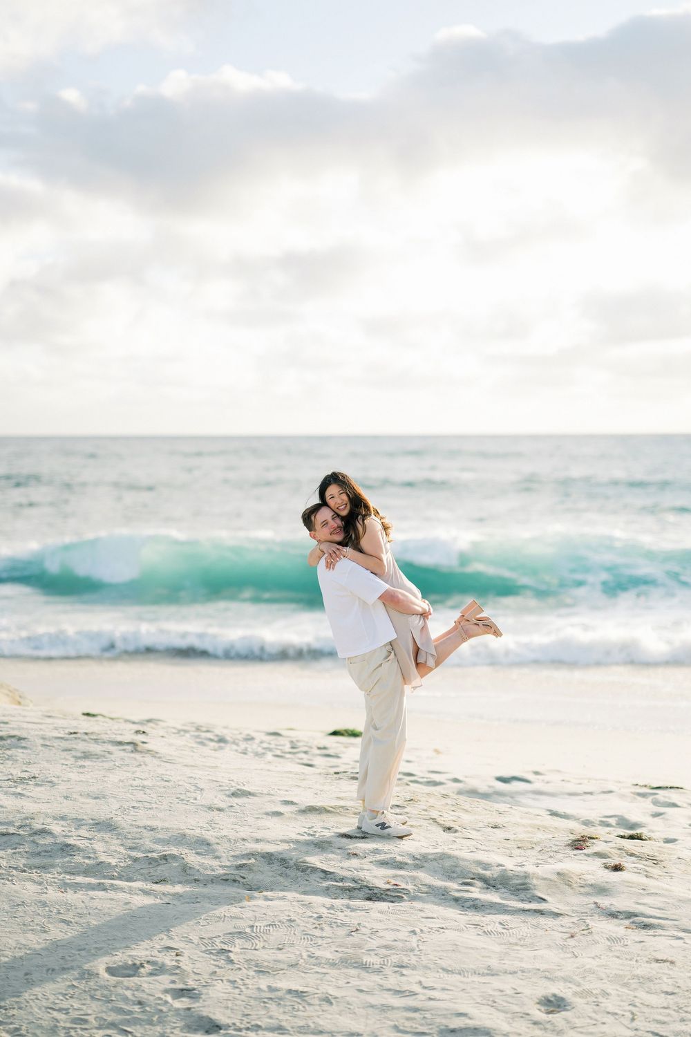 A romantic couple shares a joyful moment on a scenic beach with turquoise waves and cloudy skies in the background.
