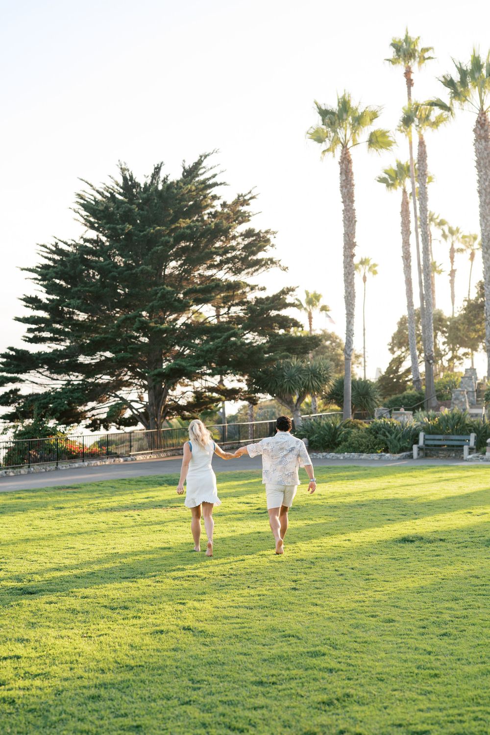A couple in white outfits holding hands while running across a sunny lawn with palm trees and cypress in the background.