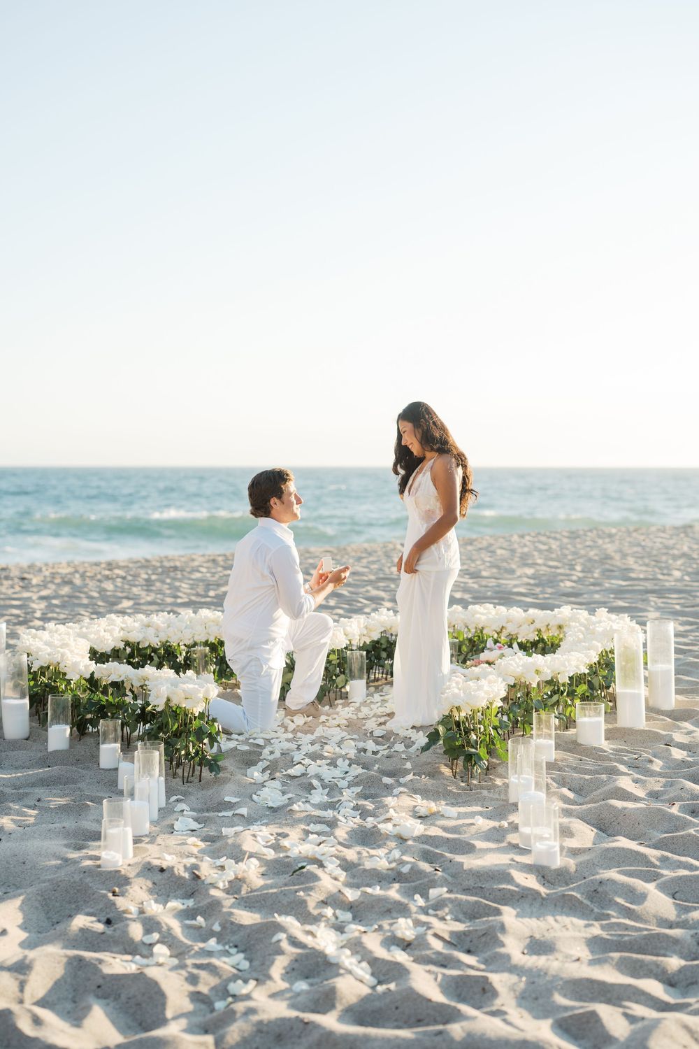 A romantic beach proposal scene with white pillars and floral decorations on the sandy shore.