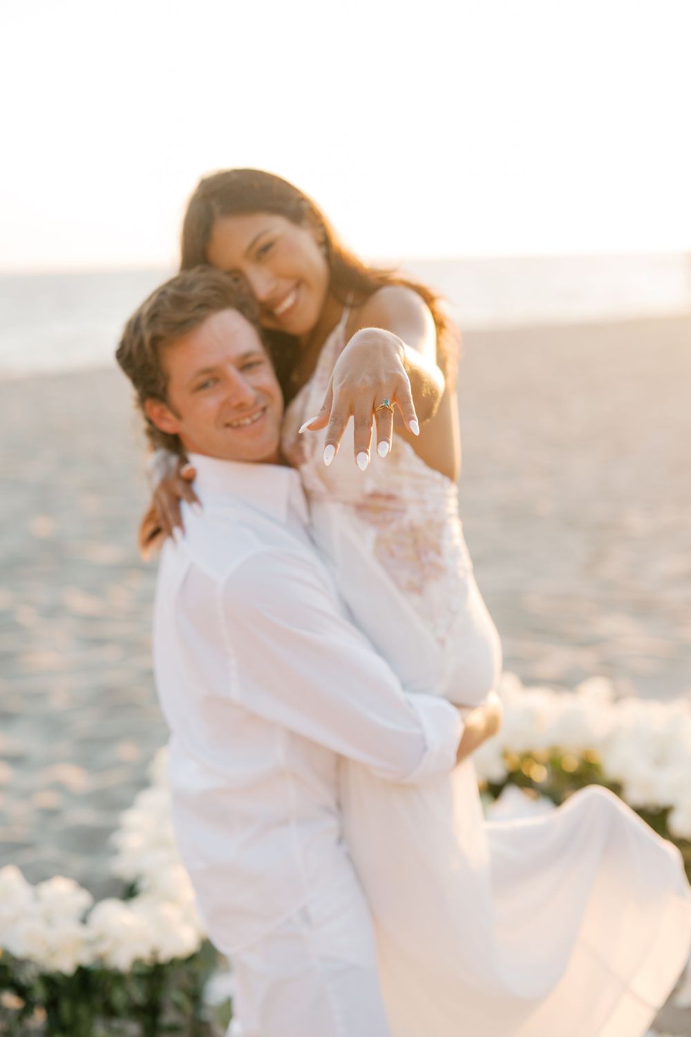 A romantic couple in white outfits embraces on a sunny beach during sunset creating an intimate moment.