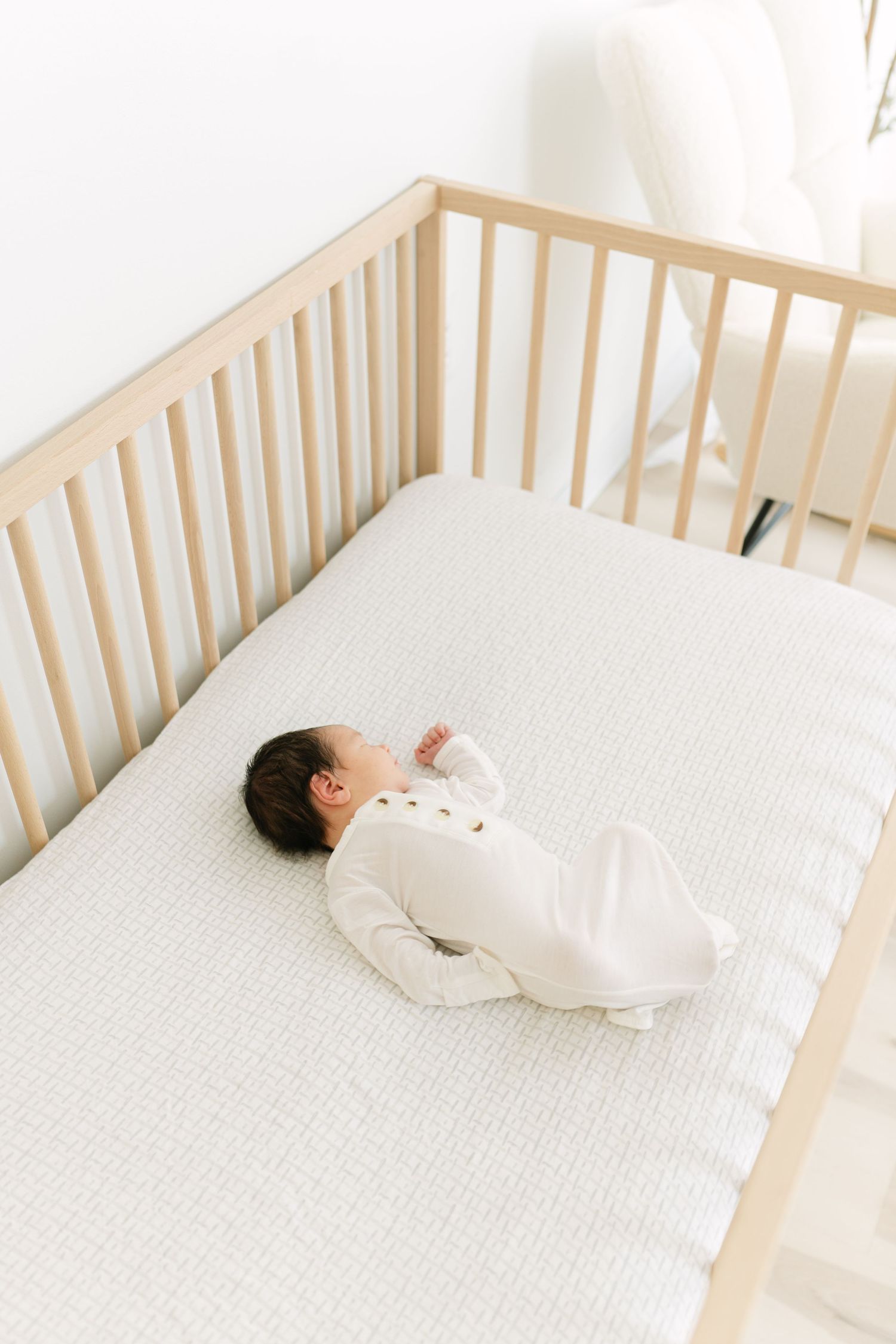 Baby in white sleeper peacefully lying in wooden crib with white bedding.