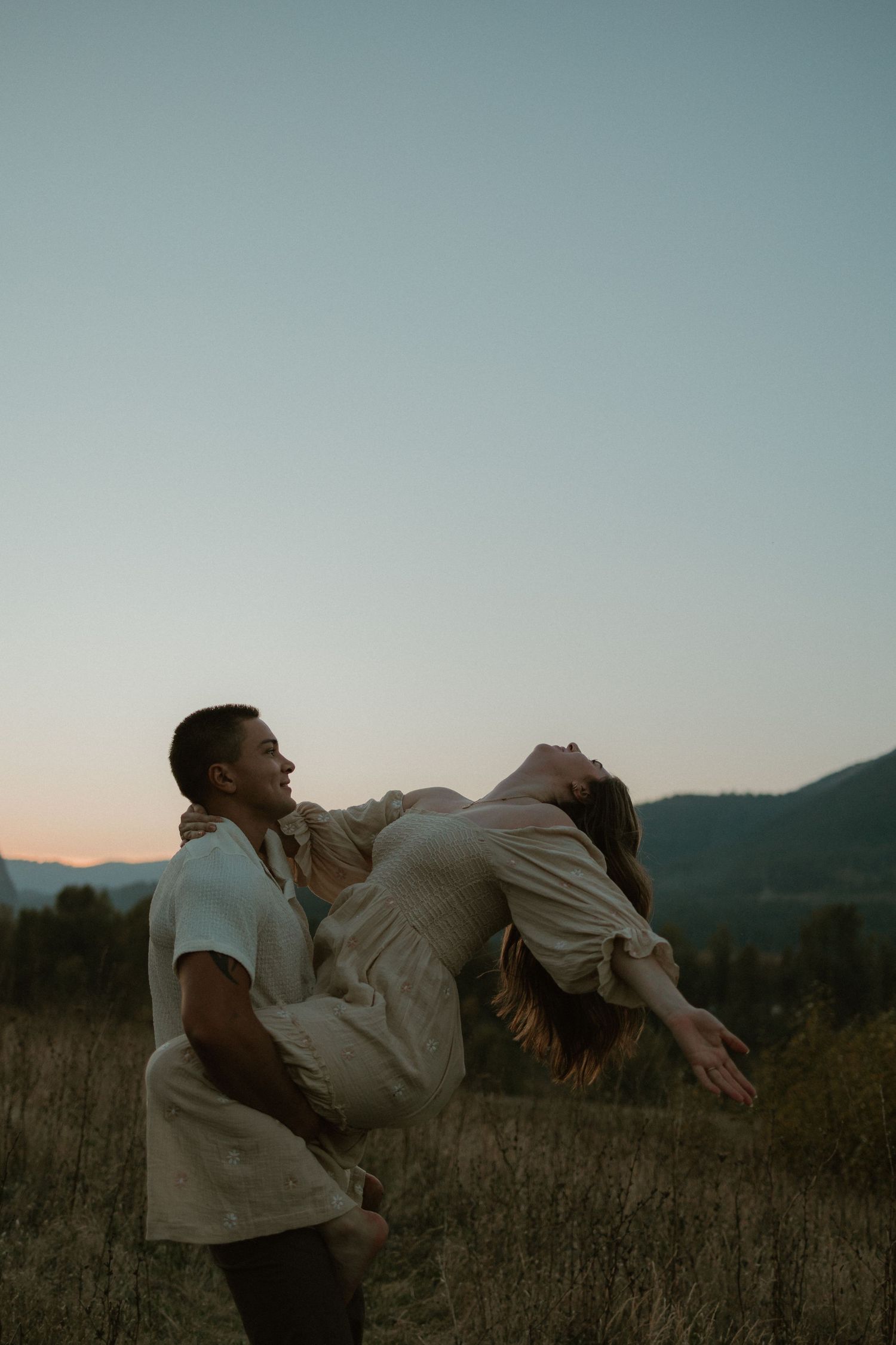 A couple shares a romantic moment in a grassy field at dusk with mountain silhouettes in the background.