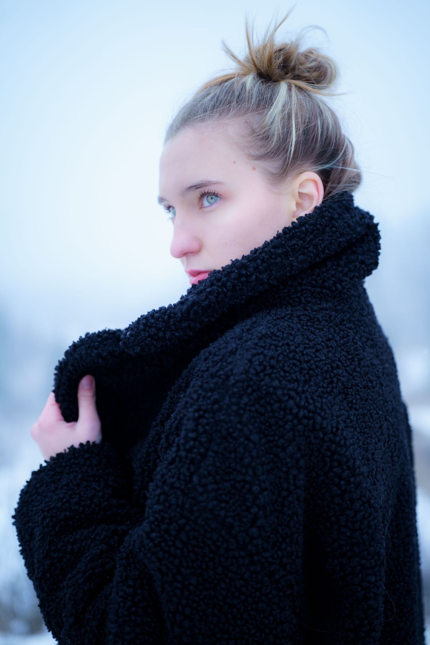 Side profile portrait in a cozy black teddy coat against a snowy winter backdrop.