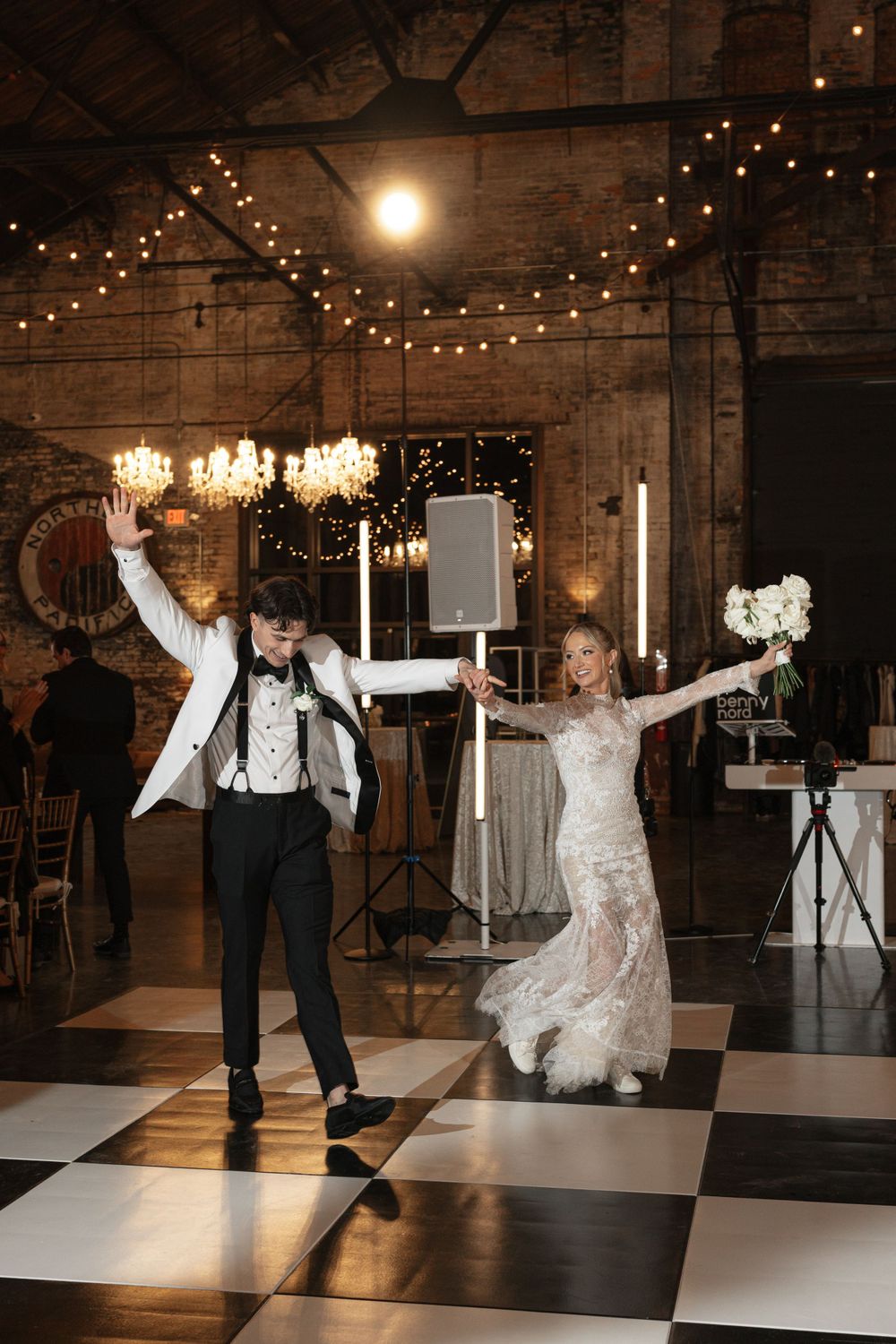 Wedding couple celebrating on checkered dance floor under string lights and chandeliers.