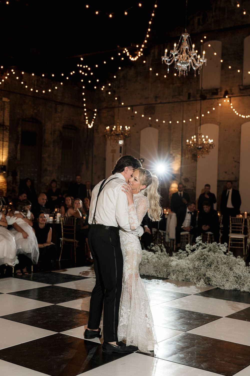 Newlyweds share an intimate slow dance moment under romantic string lights and crystal chandeliers.