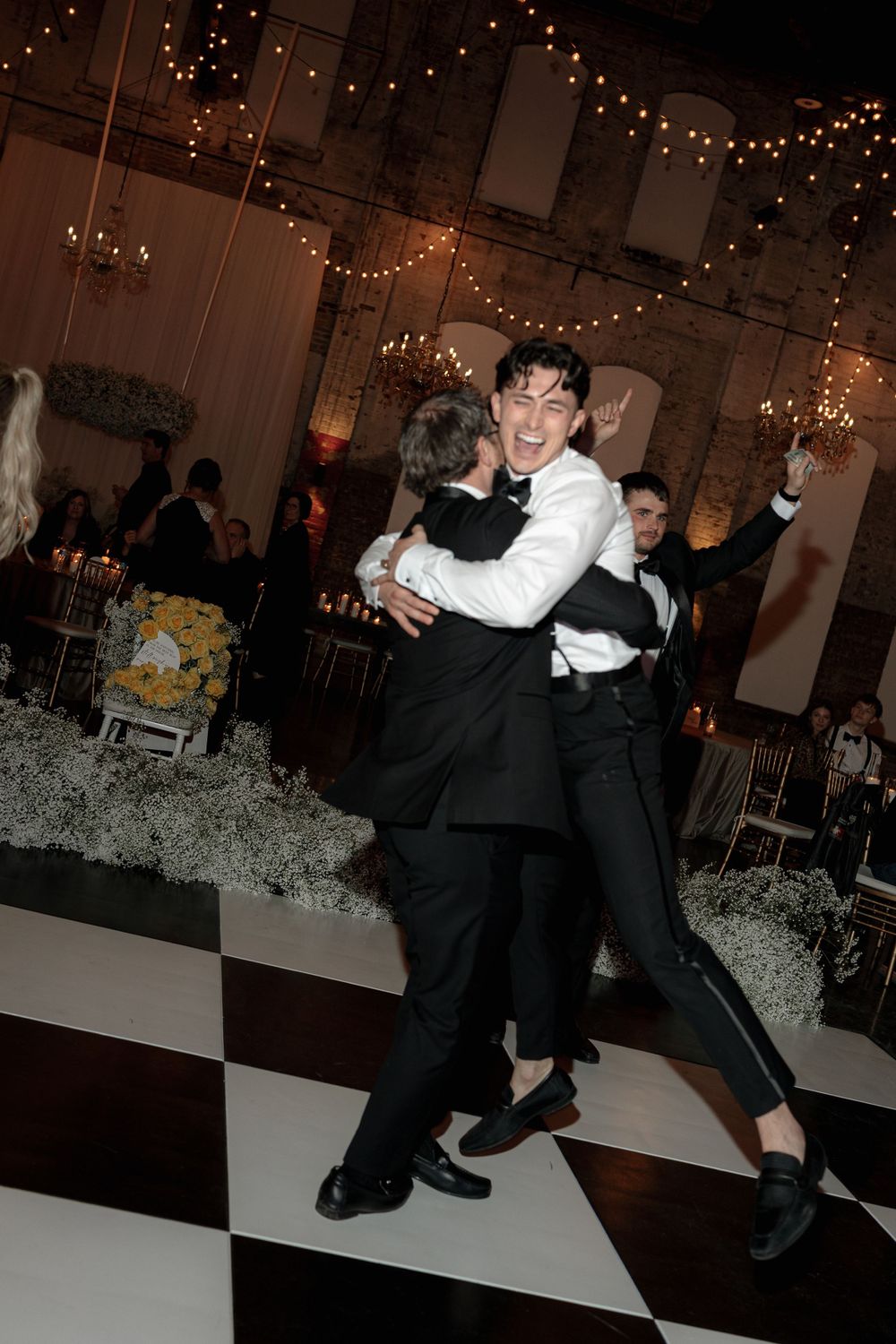 Wedding guests share a joyful dance embrace on the black and white checkered floor.