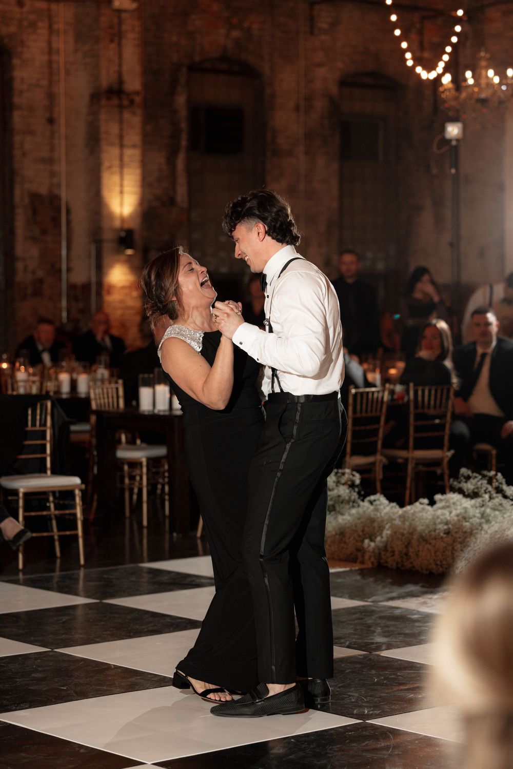 A couple shares a heartfelt dance on a checkerboard floor at an elegant reception venue with string lights overhead.